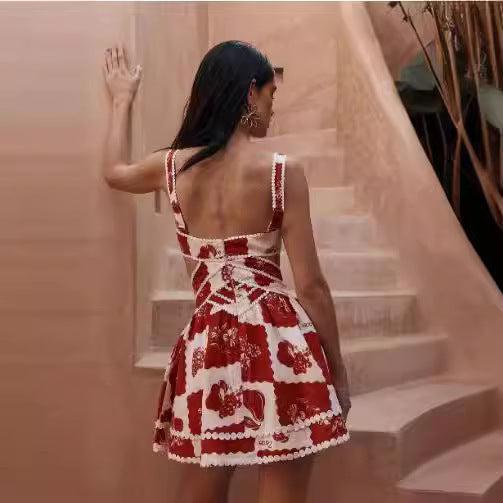 Woman wearing a red and white patchwork floral mini dress with side cutouts and wide straps, leaning against pastel stairs in a sunlit outdoor setting.