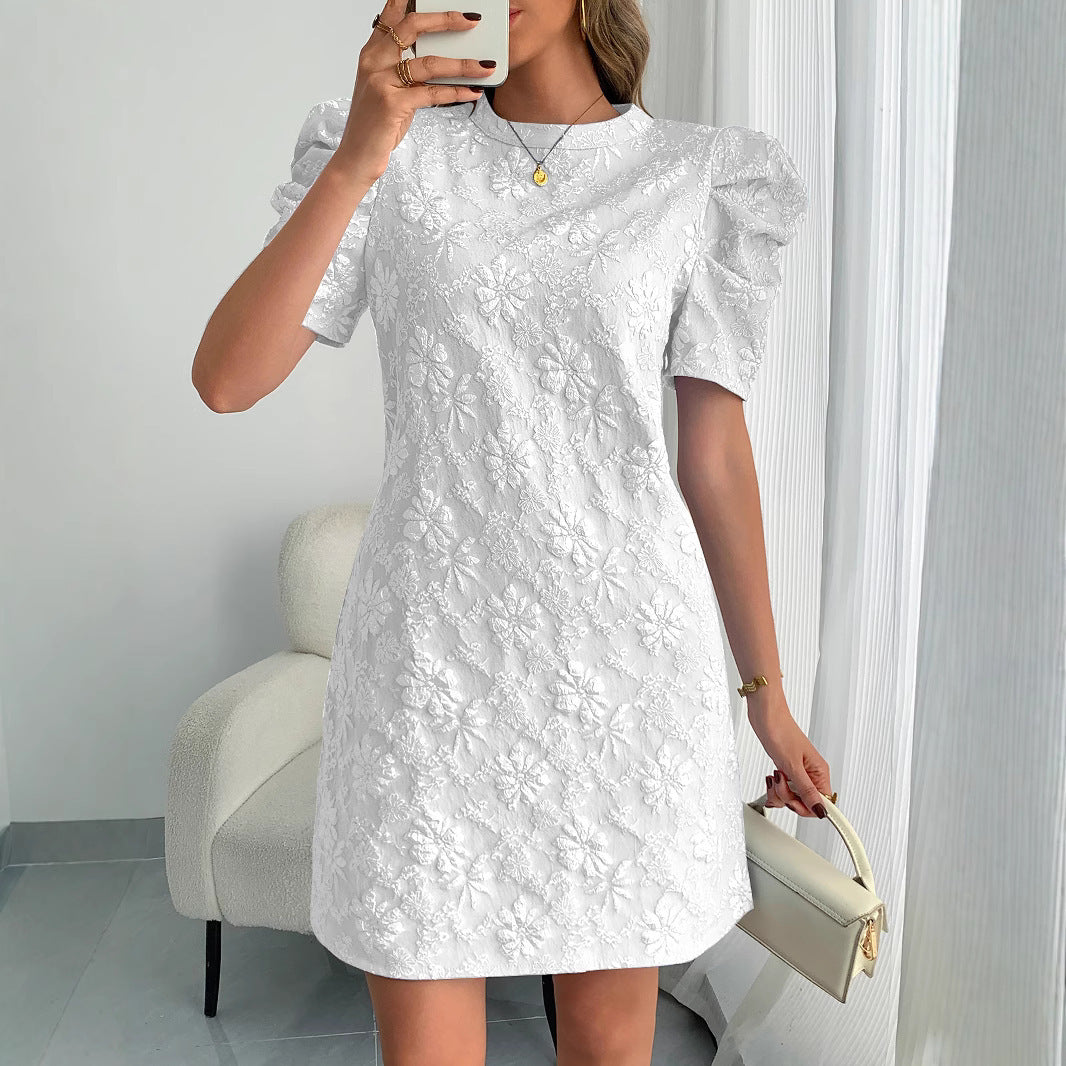 Woman wearing a soft pink mini dress with white floral embroidery and puff sleeves, holding a small white handbag against a neutral background.